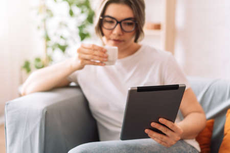 Girl drinks coffee and reads book on the tablet while sitting on the couchの写真素材