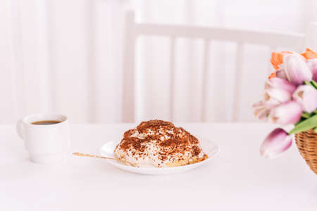 Piece of cake on white plate next to fork and cup of coffee on white table with flowersの写真素材