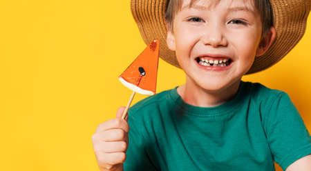 Boy in hat holding lollipop in the form of piece of watermelon in his hand against yellow backgroundの写真素材