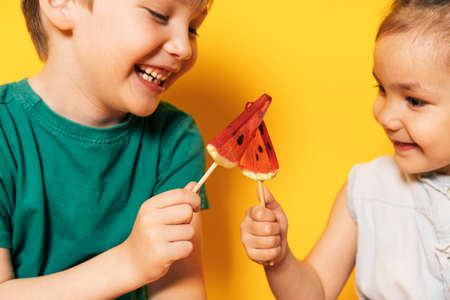 Boy with girl hold watermelon-shaped lollipops in their hands against yellow backgroundの写真素材
