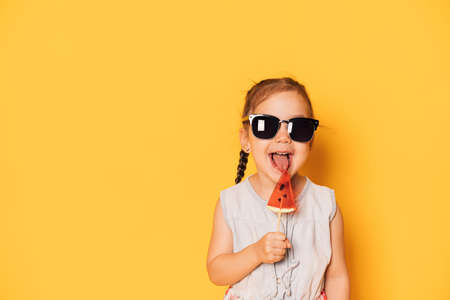 Girl in glasses licks lollipop in the form of piece of watermelon against yellow background