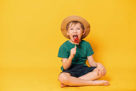 Boy in hat licks lollipop in the form of piece of watermelon while sitting on yellow backgroundの写真素材
