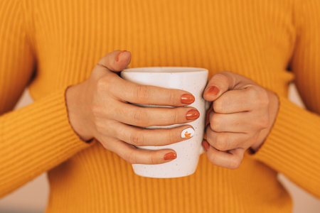 Girl in an orange sweater holding white mug with hot drink in her handsの写真素材