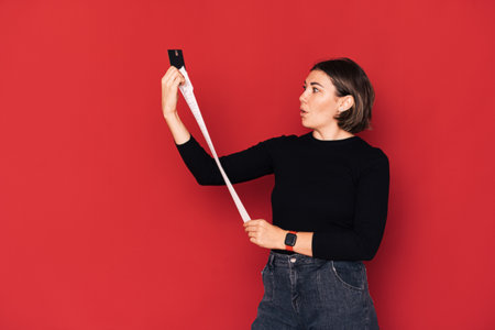 Lady looking at long shopping receipt holding credit card against red backgroundの写真素材