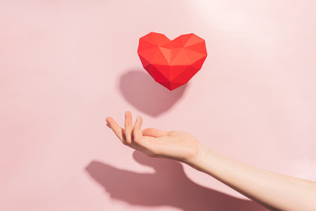 Red volumetric heart made of paper over hand with hard shadow against pink backgroundの写真素材