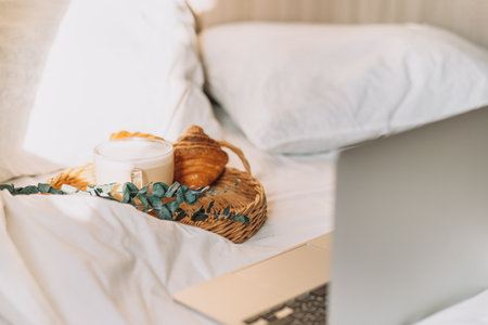 Wicker tray with croissants and coffee on white bed linen with laptopの写真素材