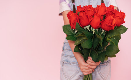 Little girl in denim overalls holding bouquet of roses behind her back against pink background, mothers dayの写真素材