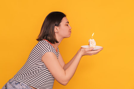 Girl in front of yellow background blows out candles on piece of cakeの写真素材