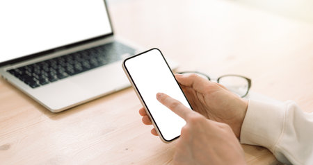 Man using smartphone in front of laptop with white screen on wooden tableの写真素材