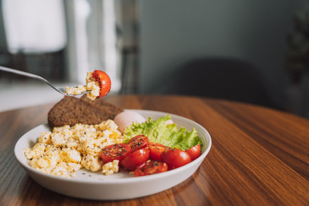 Sliced vegetables with scrambled eggs and ham on white plate with fork on wooden tableの写真素材