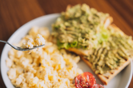 Omelet on a fork in front of a healthy breakfast with tomatoes and toast with avocado on a white round plateの写真素材