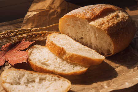 Sliced fresh loaf of bread in paper packaging with ears of wheat and maple leaves on a wooden tableの写真素材