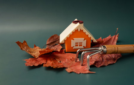 Dry maple leaves around a miniature orange house with a rake on a green backgroundの写真素材