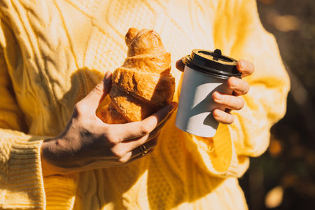 Lady in a yellow knitted sweater holding a coffee to go and a croissantの写真素材