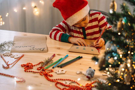 A little girl writes a letter to Santa Claus on a wooden table among pens and Christmas decorations in front of garlandsの写真素材