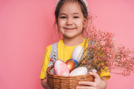 Little girl wearing bunny ears on her head holding a basket of Easter eggs in her hands against a pink backgroundの写真素材
