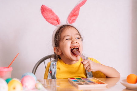 A little girl wearing bunny ears on her head eats chocolate from a spoon at a wooden table on which...