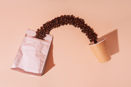 Coffee beans spill out of packaging from a paper cup on a beige backgroundの写真素材