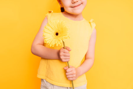 Little girl holding a yellow flower in front of a yellow backgroundの写真素材