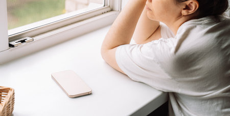 Side view of a woman daydreaming by the window with a phone lying nearbyの写真素材