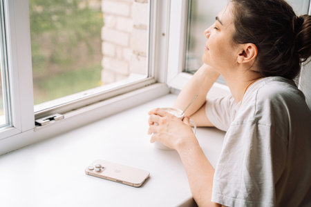 Smiling woman enjoying a hot drink by the window with a smartphone nearbyの写真素材
