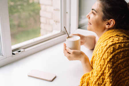 Woman smiling while holding coffee and looking out the window, enjoying tech-free timeの写真素材