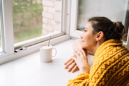 Woman relaxing with a cup of coffee near the window, wrapped in a yellow sweaterの写真素材