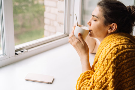 Woman sipping coffee in a knitted yellow blanket by the window, phone placed asideの写真素材