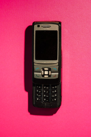 Old mobile phone with physical keypad on hot pink paper background. Flat lay with retro tech vibes.の写真素材