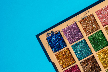 Messy glitter eyeshadow palette on bold cyan background. Imperfect beauty in vibrant pop style.