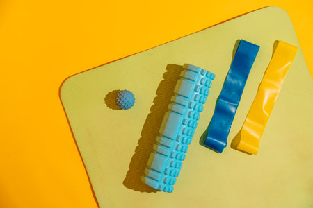 Flat lay on yoga mat with foam roller blue and yellow bands and massage ball over yellow backdrop...