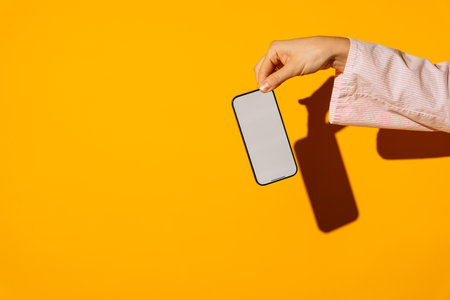 Hand holding modern smartphone with blank screen on vibrant yellow backgroundの写真素材