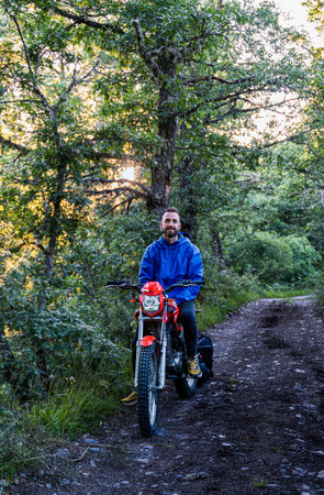 man with blue jacket sitting on a red motorbike on a path in the middle of natureの写真素材