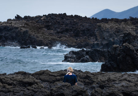 Woman in backwards with blonde hair standing on the rocks near the sea looking the wavesの写真素材