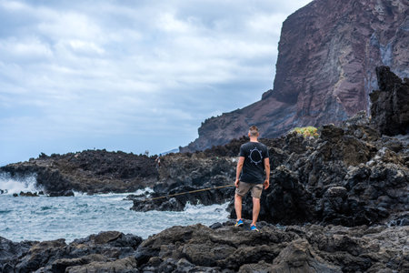 Backwards of caucasian young man standing in the black rocks near the seaの写真素材