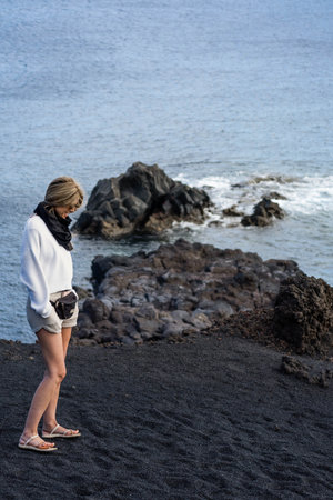 Woman with blonde hair looking at the floor on the black sand near the seaの写真素材