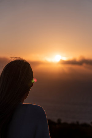 Balckwards of blonde woman in a sunset sky and sun through the clouds.Tranquil seascape and horizon over the waterの写真素材