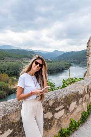 Caucasian woman with long blonde hair with her smartphone supported on the wall near de river in the villageの写真素材