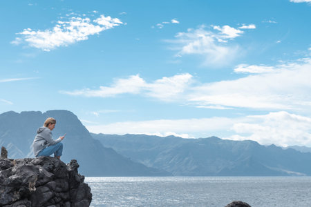 Woman sitting typing with her smartphone on the rocks seascapeの写真素材