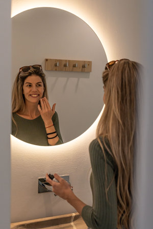 Young beauty blonde woman with long hair checking her skin at mirrorの写真素材