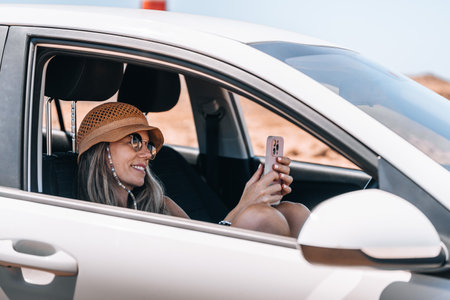 Woman with hat smiles while using her smartphone in a parked carの写真素材