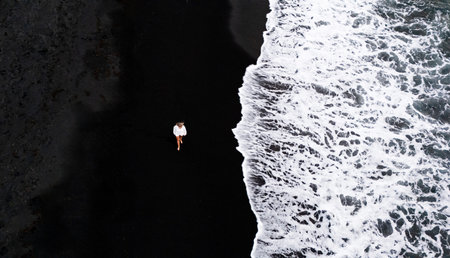 Top drone view of a woman walking along black sand at sunset near ocean waves