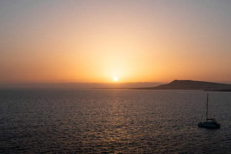 Sunset over calm waters with distant hills and a sailboat near the shoreの写真素材