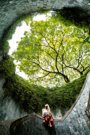 Blonde tourist sitting on spiral staircase inside fort canning park, admiring lush green treeの写真素材