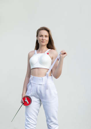 Portrait of isolated confident beautiful woman wears white fencing costume: chest protector, epee,...
