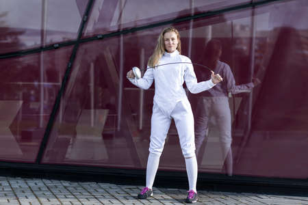 Young woman posing on red glass background, wears fencing costume: white jacket, pants, glove, socks, shoes; holds sabre (epee) in hands. Athletes, professional coach, healthy lifeの写真素材