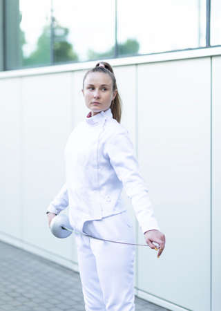Young pretty caucasian woman in white fencing costume hold epee, wears jacket, pants, looks away. Sports, healthy lifestyle, professional coach. Vertical plane.の写真素材