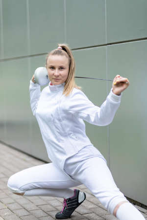 Young European woman posing in white fencing costume, holding epee on back. Vertical Expensive...