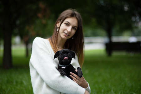 Black cute pet pug-dog of breed 'Petit Brabancon Brussels Griffon' sits on the hands of out of focus brunette in green park. Friendly social companion. Summertime. Horizontal planeの写真素材