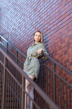 Young lovely European slav female blondie puts her hand on handrail standing on stairs, red bricks wall of building is on background, wears green - khaki denim Relaxed in summer daの写真素材